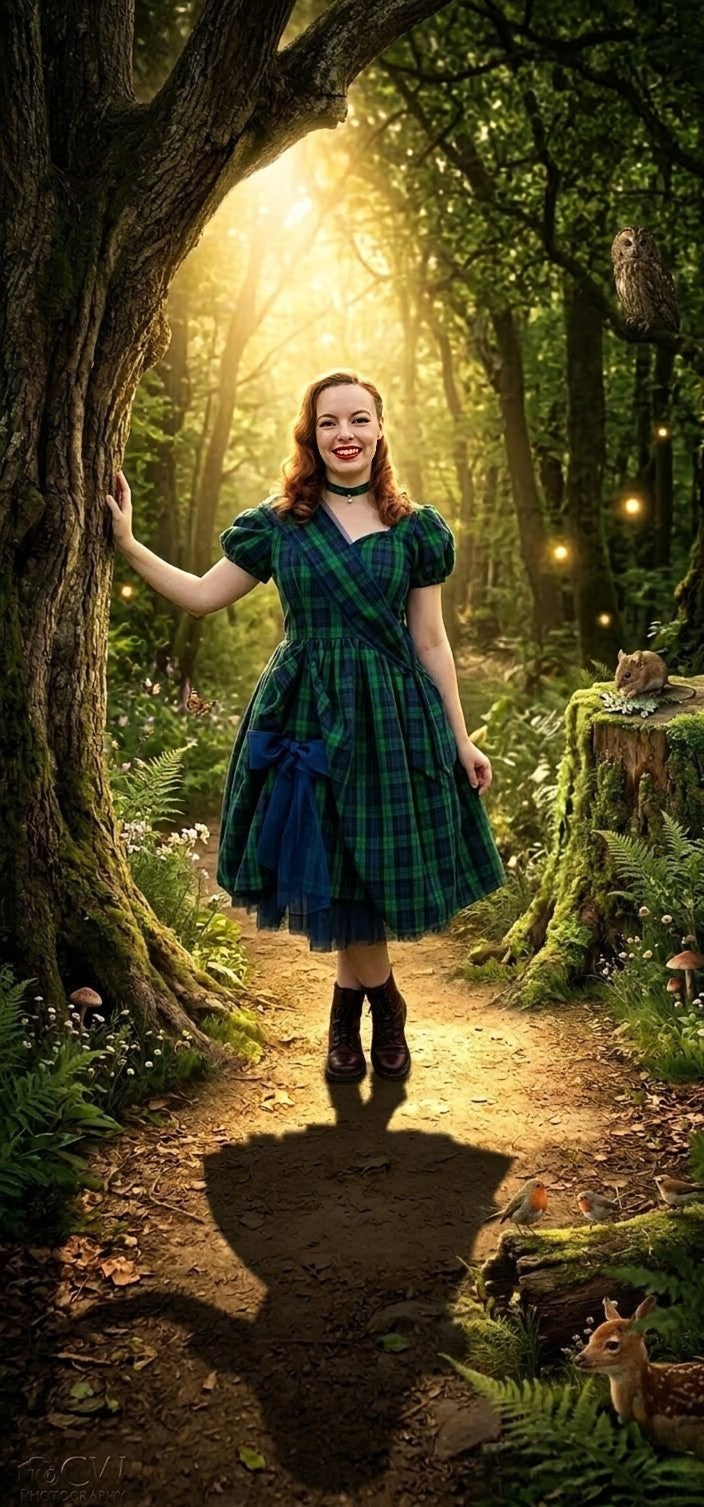 A photo of a woman in an elegant emerald green and blue plaid tartan gown standing in an enchanted, sun-dappled forest path.