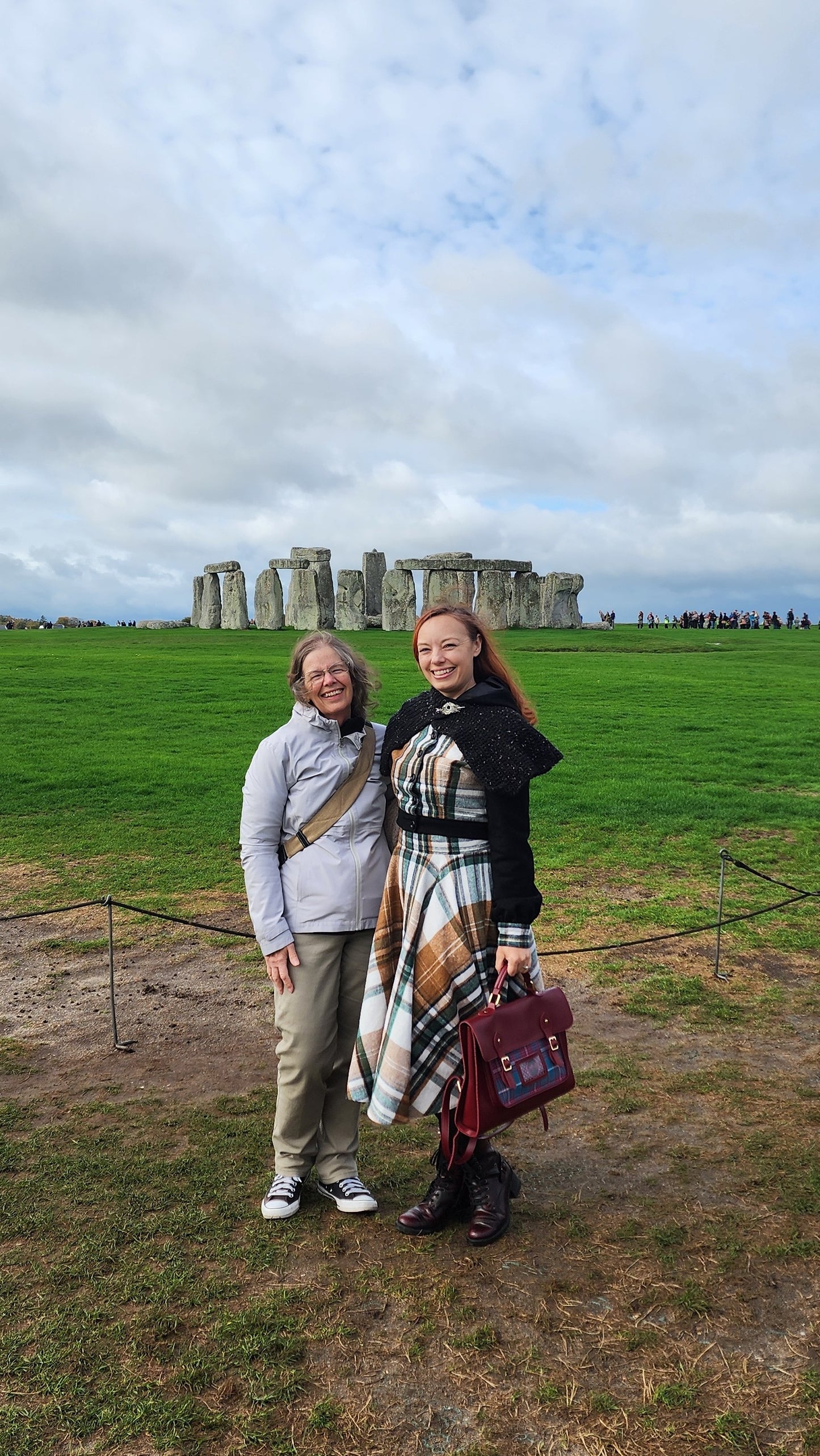 A woman standing in front of the Stonehenge stone circle wearing a vintage-inspired brown and green plaid flannel circle skirt and a matching jacket with black wool sleeves. The wind is catching the full sweep of the skirt, showing its movement and volume against the historic landscape.