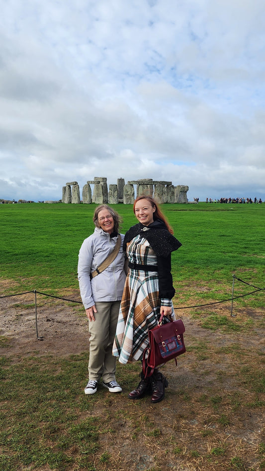 A woman standing in front of the Stonehenge stone circle wearing a vintage-inspired brown and green plaid flannel circle skirt and a matching jacket with black wool sleeves. The wind is catching the full sweep of the skirt, showing its movement and volume against the historic landscape.