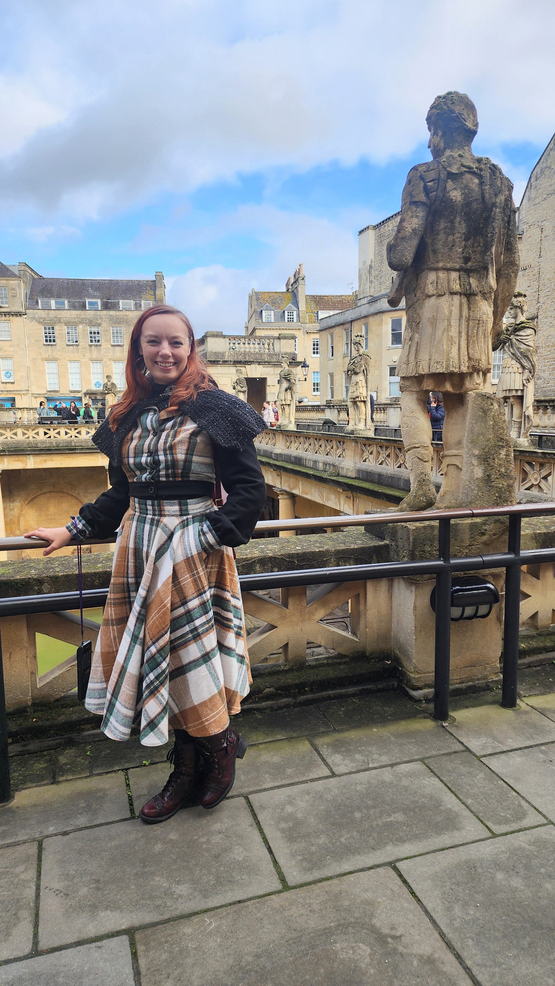 A medium shot of a woman at the Roman Baths in Bath, England, wearing a tailored plaid bodice with black wool sleeves and a hood. The image highlights the structured fit of the jacket and the traditional button details, set against ancient stone pillars.