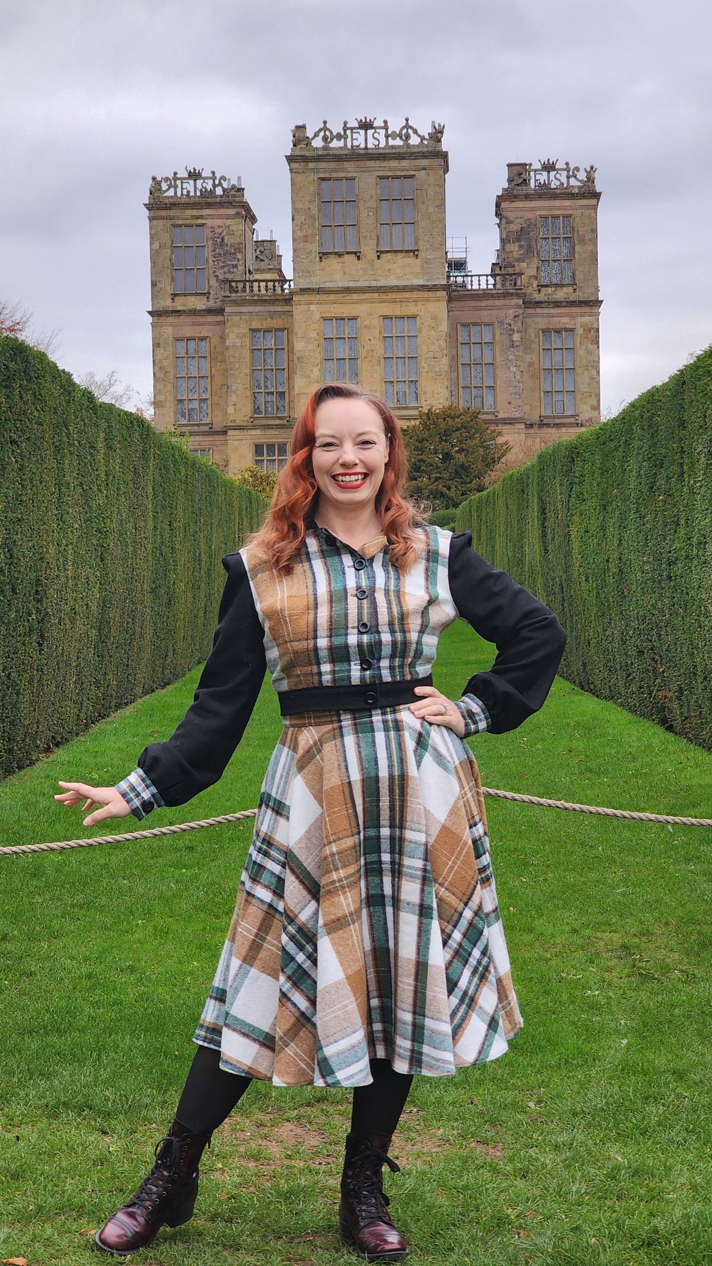 A full-length portrait of a woman in a plaid flannel travel set standing before the grand facade of Hardwick Hall in Derbyshire, England. The outfit features a high-waisted circle skirt and a hooded jacket, blending historical 16th-century silhouettes with modern textiles.