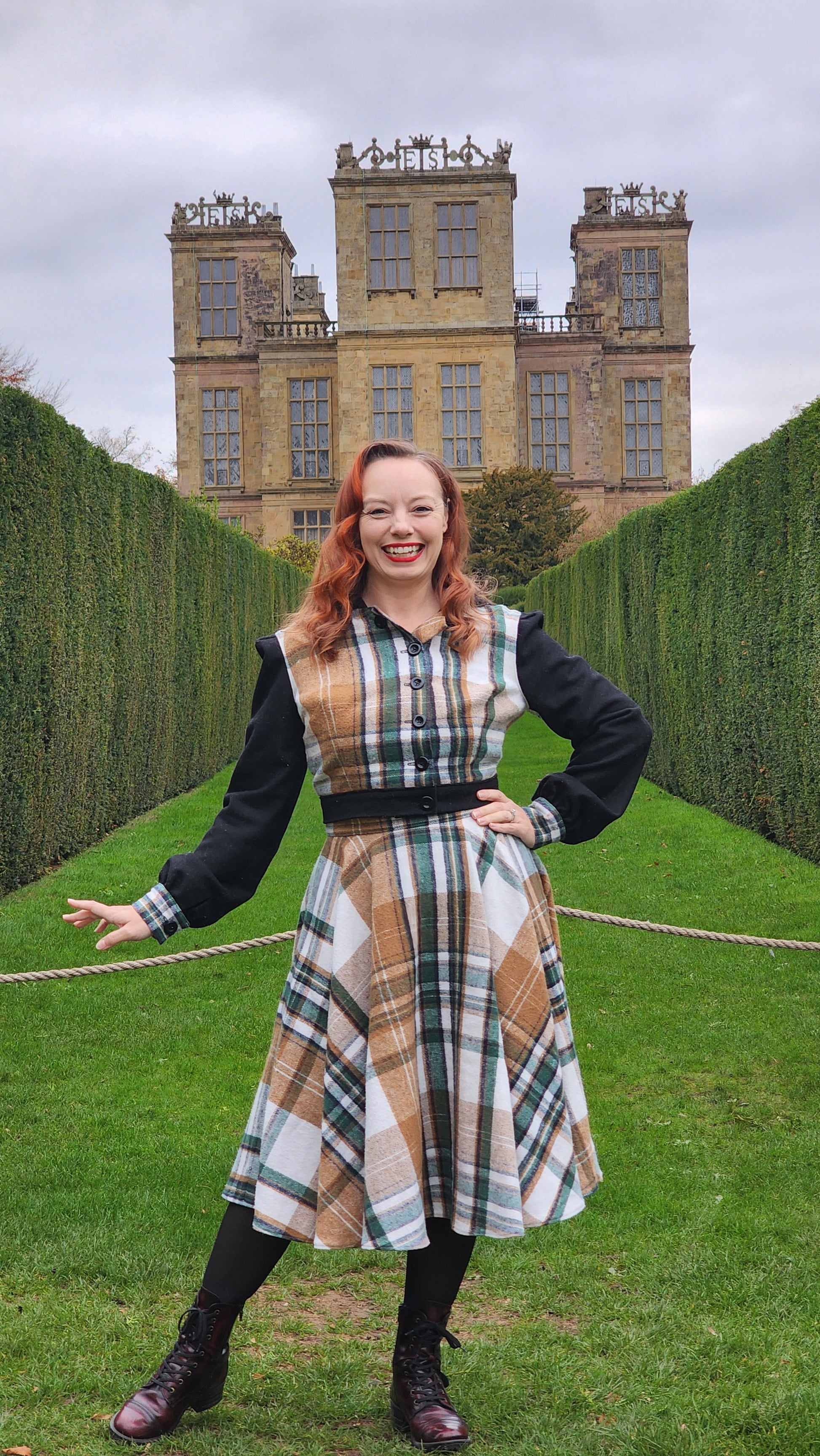 A full-length portrait of a woman in a plaid flannel travel set standing before the grand facade of Hardwick Hall in Derbyshire, England. The outfit features a high-waisted circle skirt and a hooded jacket, blending historical 16th-century silhouettes with modern textiles.