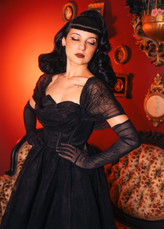 Woman in a black vintage inspired spiderweb pinuo dress and gloves posing against an ornate wall. 