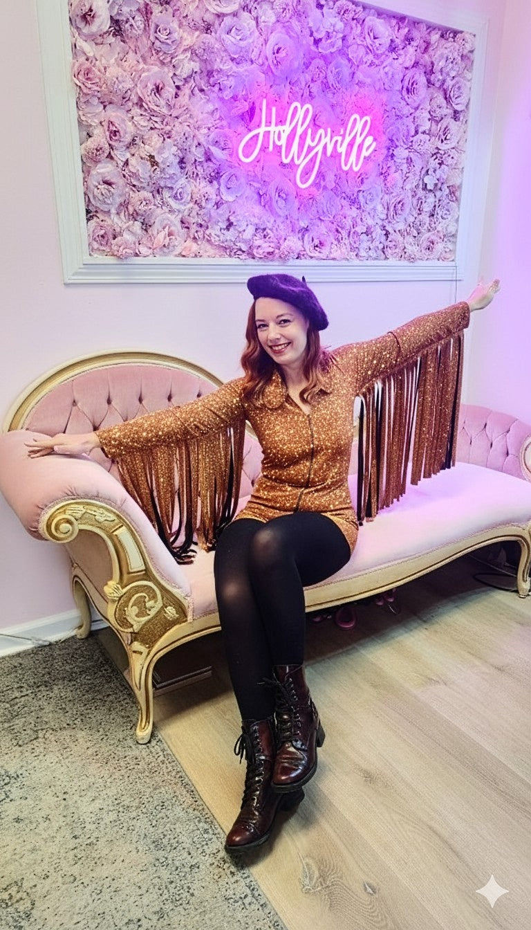 Woman in a 1970's vintage inspired gold fringe sequin romper sitting on a pink couch with a 'Hollyville' sign in the background
