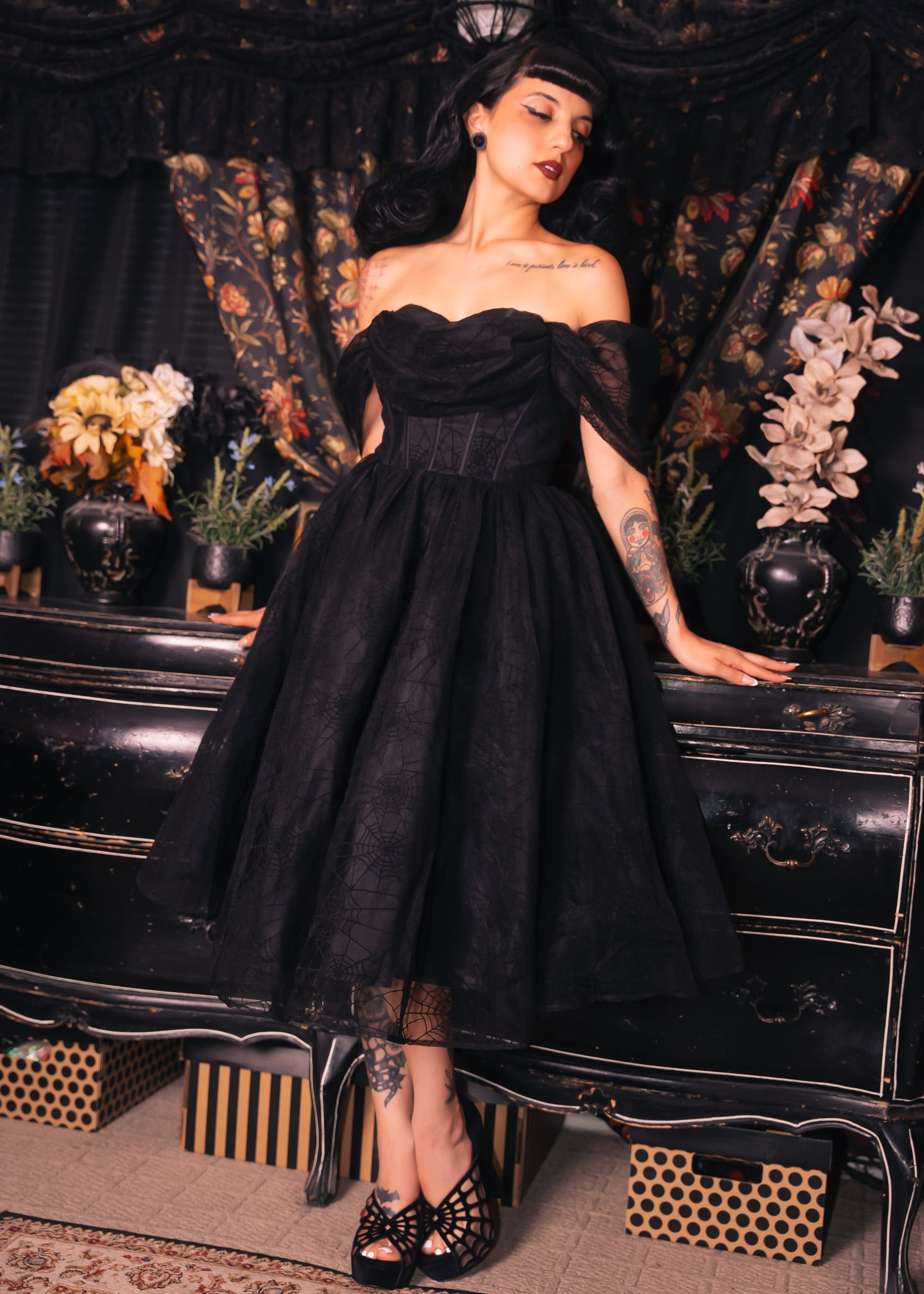 Woman in a black Halloween vintage inspired spiderweb dress standing in front of a decorative backdrop with flowers and furniture.  